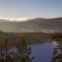 Snødekte trær skuer ut over en frossen innsjø ved soloppgang. Det myke sollyset filtreres gjennom tåke og skyer og kaster et varmt, gyllent skjær over de skogkledde åsene og skaper et fredfylt vinterlandskap.
